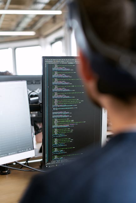 A close-up view of a laptop computer displaying lines of code on the screen, with a focus on programming syntax such as variables, functions, and comments. The laptop is situated on a desk with a dark